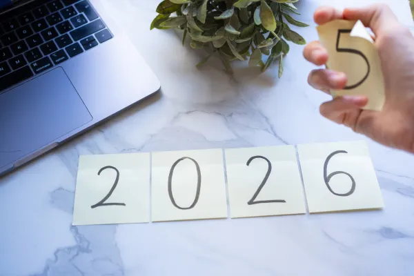 Hand holding a sticky note with the number 5, with sticky notes displaying "2026" on a desk.