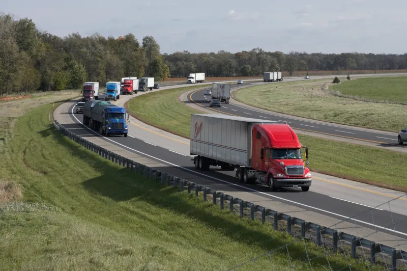 Red and white semi-truck driving on a curved highway with cars in the background.
